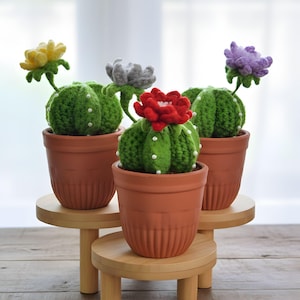 Puede incluir: Tres cactus de ganchillo hechos a mano con flores coloridas en macetas de terracota. Los cactus son verdes con detalles de cuentas blancas, y las flores son amarillas, grises, rojas y moradas. Las plantas están sobre pequeños soportes redondos de madera.