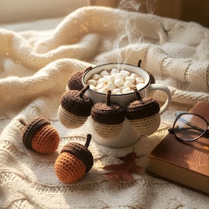 Può includere: Una scena accogliente con una tazza bianca piena di cioccolata calda e marshmallow, circondata da ghiande all'uncinetto nei toni del marrone e dell'arancione. Una calda coperta di maglia color crema e un libro rilegato in pelle completano l'ambientazione autunnale.