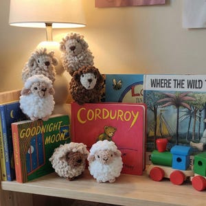 May include: Handmade crochet sheep figurines in white, beige, and brown are arranged with children's books such as "Corduroy" and "Goodnight Moon" on a wooden shelf. A toy train is also present in the image.