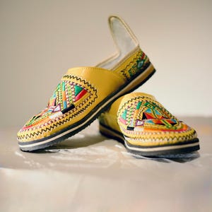 Puede incluir: Un par de zapatos sin cordones de cuero amarillo con coloridos patrones bordados en la parte superior. Los zapatos tienen una suela negra y un acento blanco en el talón. El bordado presenta diseños geométricos en rojo, verde, azul y naranja.