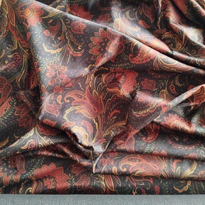 May include: Dark fabric with a paisley pattern in shades of red, orange, and gold against a black background. The fabric is folded, showing the intricate design and texture. A roll of dark gray fabric is visible at the bottom.
