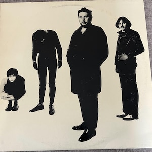 May include: A square vinyl record cover featuring a black and white photograph of four individuals against a plain off-white background. One person is crouching, one is standing with head bowed, and two are standing upright in dark clothing.