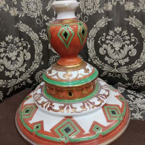 May include: A decorative ceramic pedestal with a tiered design. The base is white with red and green geometric patterns, accented with gold trim. The central section is orange with green and black diamond shapes. The top is white.