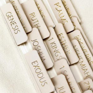 May include: A set of beige adhesive bible index tabs featuring gold foil lettering. The tabs are arranged in a staggered pattern, displaying book names including Genesis, Exodus, Joshua, Ruth, 1 Kings, Esther, Psalms, Nehemiah, and Proverbs in a clear font.