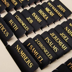 May include: A collection of black rectangular Bible index tabs featuring gold foil lettering. Each tab displays the name of a specific book of the Bible, including Numbers, 1 Samuel, 2 Chronicles, Psalms, Jeremiah, Joel, Nahum, Ruth, 1 Chronicles, Job, Isaiah, Hosea, Micah, Zechariah, and John.