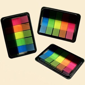 May include: Three black plastic dispensers containing stacks of translucent adhesive page markers in neon colors including pink, orange, yellow, green, and blue. Each rectangular dispenser holds five distinct rows of colorful sticky index tabs.