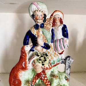 Pode incluir: Estatueta de cerâmica vintage que retrata um homem, uma criança e uma raposa em uma cena campestre. O homem usa um casaco azul e um chapéu grande. A criança usa uma camisa branca e um casaco azul. A raposa é vermelha.