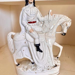 Op de afbeelding: Een witte keramieken beeldje dat een persoon in historische kleding op een wit paard afbeeldt. De figuur draagt een witte hoed, jas en broek, met een rode riem. Het paard heeft gouden accenten en zwarte hoeven.