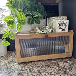 May include: A bamboo bread box with a clear, ribbed glass door sits on a countertop. A white ceramic pot with a green plant, a decorative box with lemons, and a sign that says "I just want to SIP COFFEE & pet my DOG" are on top.