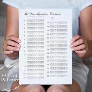 Op de afbeelding: Een wit papier met de titel "30 Day Hydration Challenge" wordt door een persoon vastgehouden. Het papier heeft een raster met vakjes om de waterinname bij te houden. De tekst "A5, A4, US Letter" en "direct downloaden" zijn ook zichtbaar.