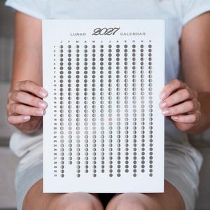 Pode incluir: Um calendário lunar de papel branco para 2021, segurado por uma pessoa. O calendário apresenta uma série de círculos perfurados que representam as fases lunares. A palavra "LUNAR" está no topo, com o ano "2021" em uma fonte estilizada. Os meses são abreviados no topo.