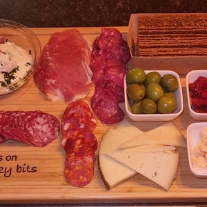May include: A wooden charcuterie board with various meats, cheeses, olives, and crackers. The board includes salami, prosciutto, and cheese slices. Small white bowls hold olives, sun-dried tomatoes, and cheese cubes. The text "Off my tits on picky bits" is engraved on the board.
