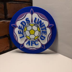 Puede incluir: Emblema redondo azul del Leeds United AFC. El diseño presenta una flor blanca y morada con un balón de fútbol en el centro. Las palabras "LEEDS UNITED AFC" están escritas alrededor de la flor en blanco.