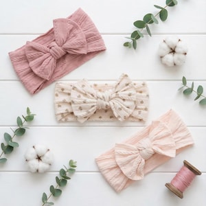 Puede incluir: Tres diademas en tonos rosa y crema se exhiben sobre una superficie de madera blanca. Las diademas presentan un diseño de lazo. Hojas de eucalipto y motas de algodón se disponen alrededor de las diademas, junto con una bobina de hilo rosa.