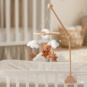 May include: A wooden baby mobile with a teddy bear, clouds, stars, and a crescent moon. The mobile is suspended from a wooden arm attached to a white crib. The bear is brown, the clouds are white, and the stars are brown.
