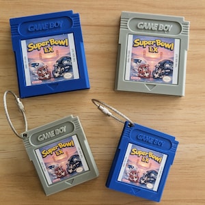 Może przedstawiać: Cztery kartridże do Game Boy w kolorze niebieskim i szarym, każdy z etykietą "Super Bowl LX" z grafiką hełmów do futbolu amerykańskiego. Kartridże mają dołączone metalowe breloczki. Tekst "GAME BOY" jest widoczny na każdym kartridżu.