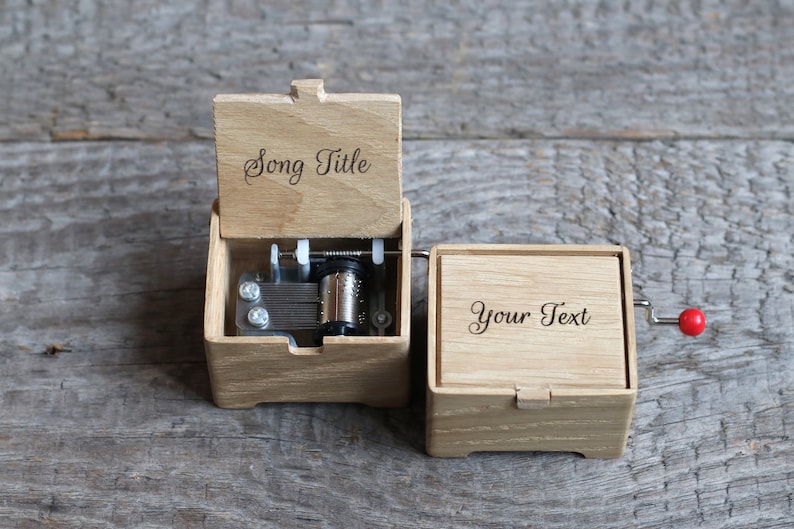May include: A wooden music box with a red crank. The top of the box is open and shows the inner workings of the music box. The top of the box is engraved with the words "Song Title". The bottom of the box is engraved with the words "Your Text".