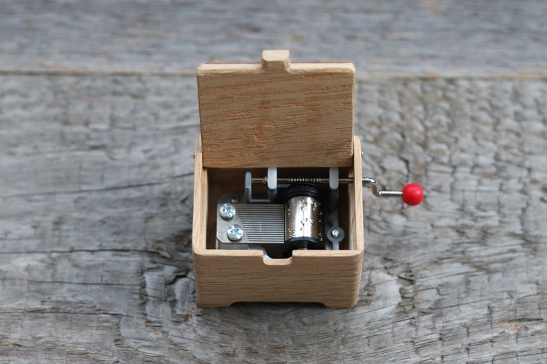 May include: A wooden music box with a red crank and a silver winding mechanism. The lid is open, revealing the inside of the box.