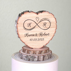 Könnte beinhalten: Holz-Tortenaufsatz in Herzform mit einem Unendlichkeitszeichen und den Initialen "K" und "R" graviert. Der Text "Karen & Robert 10.03.2025" ist ebenfalls auf dem Aufsatz graviert.