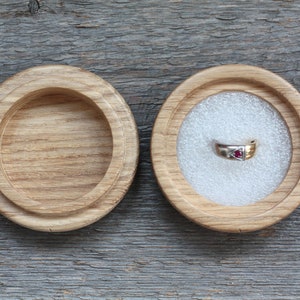 Puede incluir: Una caja de madera para anillos con tapa, abierta para revelar un interior de terciopelo blanco. Un anillo de oro con una piedra preciosa roja est&aacute; dentro de la caja.
