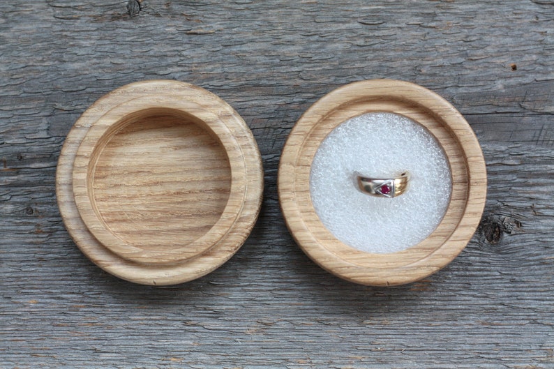 Puede incluir: Una caja de madera para anillos con tapa, abierta para revelar un interior de terciopelo blanco. Un anillo de oro con una piedra preciosa roja est&aacute; dentro de la caja.