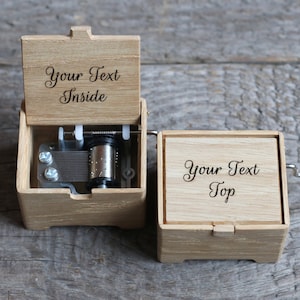 May include: A wooden music box with a hand crank on the side. The top of the box is engraved with the text "Your Text Top". The lid of the box is open, revealing the music mechanism and the text "Your Text Inside" engraved on the inside of the lid.