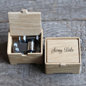 May include: A wooden music box with a red crank handle. The lid is open, revealing the inner workings of the music box. The box is engraved with the words "Song Title".