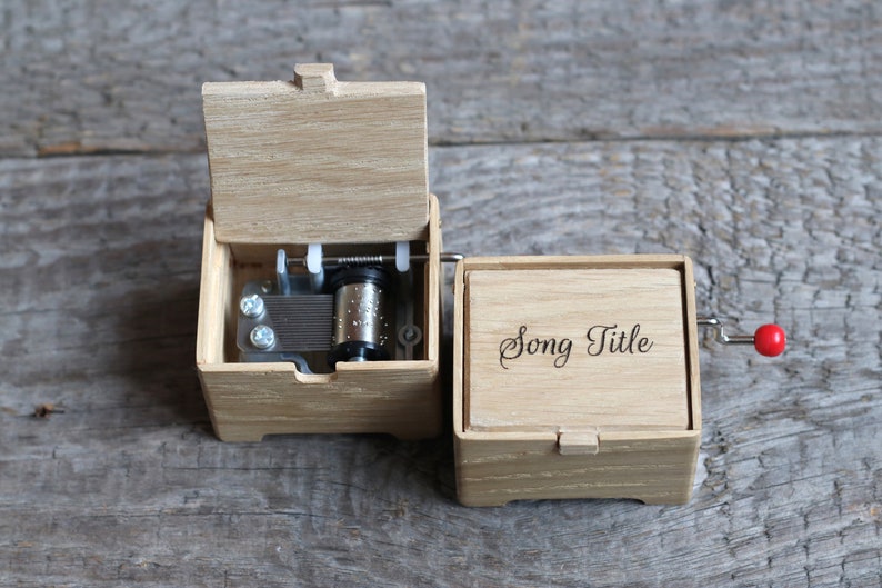May include: A wooden music box with a red crank handle. The lid is open, revealing the inner workings of the music box. The box is engraved with the words "Song Title".