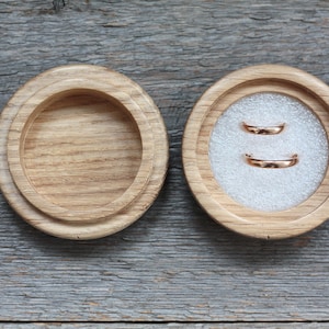 Puede incluir: Dos alianzas de oro en una caja de anillos de madera con un interior de terciopelo blanco.
