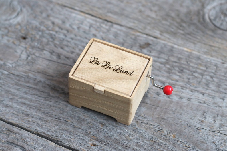 May include: A wooden music box with a hand crank and a red knob. The lid is engraved with the words "La La Land".