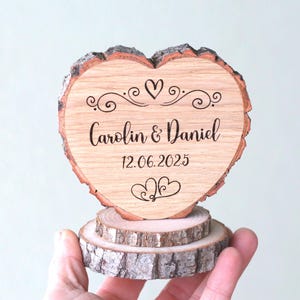 Op de afbeelding: Een houten hartvormige taarttopper voor een bruiloft. De topper is gegraveerd met de namen "Carolin & Daniel" en de datum "12.06.2025". Het hart staat op een basis van twee kleine houten schijfjes. De randen van het hart en de basis hebben een natuurlijke schorsafwerking.
