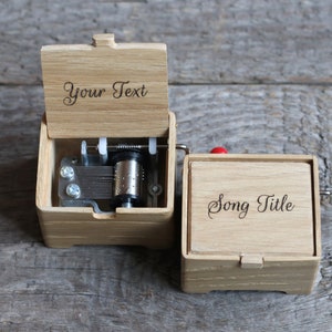 May include: A wooden music box with a hand crank. The top of the box is open and the inside mechanism is visible. The box has a personalized inscription that says "Your Text" and the bottom of the box has an inscription that says "Song Title".