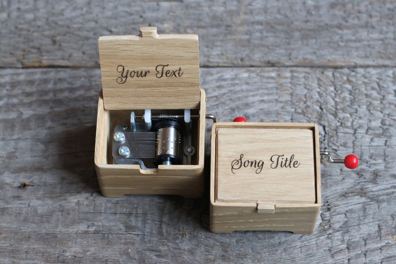 May include: A wooden music box with a hand crank. The top of the box is open and the inside mechanism is visible. The box has a personalized inscription that says "Your Text" and the bottom of the box has an inscription that says "Song Title".
