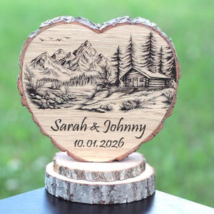 Op de afbeelding: Een hartvormige houten schijf met een brandmerk-landschap van bergen, dennenbomen en een blokhut. De tekst Sarah & Johnny en de datum 10.01.2026 zijn onder de afbeelding gegraveerd. De plaat rust op twee gestapelde, ronde houten voetstukken.