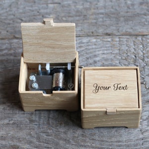May include: A wooden music box with a crank on the side. The lid is open, revealing the inner workings of the music box. The box has a blank space on the front where you can add your own text.