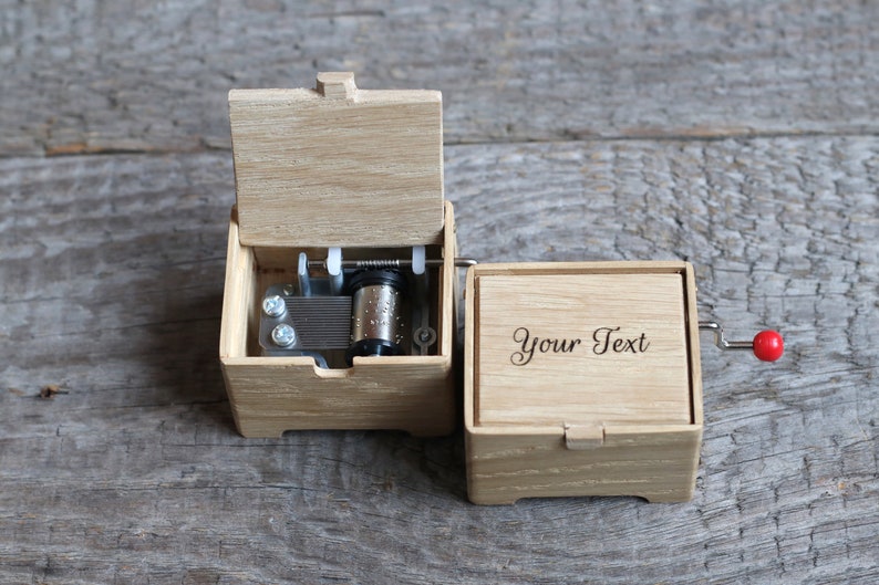 May include: A wooden music box with a crank on the side. The lid is open, revealing the inner workings of the music box. The box has a blank space on the front where you can add your own text.
