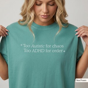 Op de afbeelding: Een zeeschuimgroen t-shirt met korte mouwen en de witte tekst "Too Autistic for chaos Too ADHD for order" in het midden op de borst, geflankeerd door kleine vierpuntige sterpictogrammen aan de linker- en rechterkant van de tekst.