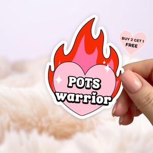 May include: A die-cut sticker featuring a light pink heart shape surrounded by bright red flames. The text "POTS warrior" is printed in bold black letters across the centre of the heart, accented by small white sparkles. A small pink heart-shaped tag in the corner displays the text "BUY 2 GET 1 FREE".