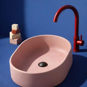 May include: A modern, oval-shaped pink bathroom sink with a black drain and a red faucet. The faucet has a curved spout and a single handle. A stack of colorful wooden blocks sits to the left of the sink. The background is a solid blue.