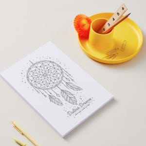 May include: A white notepad featuring a black line drawing of a dreamcatcher with feathers and the text "Deliver Dreams where your worlds are hope". A yellow desk organizer holds a wooden ruler showing 6 inches and a pencil, with paper clips.