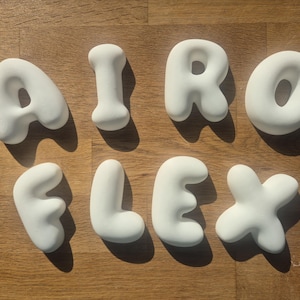 Könnte beinhalten: Ein Set aus acht einzelnen weißen, abgerundeten, dreidimensionalen Buchstaben, die das Wort AIRO FLEX bilden. Jeder Buchstabe hat eine glatte, matte Oberfläche und wirft einen deutlichen Schatten, was einen erhabenen, blasenartigen Effekt erzeugt.