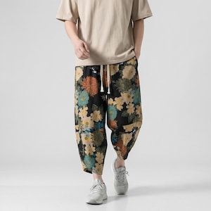 May include: Black floral print trousers with a drawstring waist. The trousers feature a colourful pattern of yellow, orange, and teal flowers. The person is wearing a beige short-sleeve shirt and grey trainers.