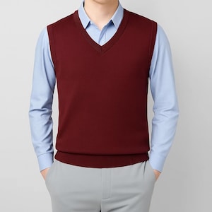 May include: A burgundy V-neck sweater vest worn over a light blue shirt. The vest has a ribbed hem and a classic fit. The model is wearing grey trousers. A classic, preppy style.