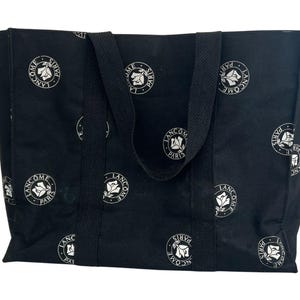 Puede incluir: Un bolso tote negro con un patrón repetitivo de logotipos circulares blancos. Cada emblema circular contiene un gráfico de rosa estilizado y el texto LANCOME PARIS. El bolso tiene dos asas de tela negra unidas a los paneles frontal y trasero.