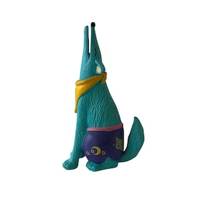 May include: A turquoise howling wolf figurine with a yellow bandana and purple shorts decorated with moons and stars. The wolf is sitting upright with its head and snout pointed upwards. The figurine has a textured surface.