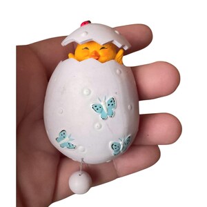 Puede incluir: Adorno blanco en forma de huevo con diseños de mariposas azul claro y un pequeño pollito amarillo que emerge de la parte superior. Una pequeña mariquita roja se posa sobre la cáscara del huevo. Una bola blanca cuelga de una cuerda en la parte inferior.
