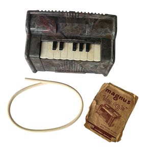 Puede incluir: Un órgano de boca vintage Magnus con carcasa de plástico veteado en gris oscuro y marrón, teclas blancas y negras, un tubo de plástico blanco flexible y un folleto de instrucciones de papel desgastado titulado Magnus blow organ instruction method and song folio.