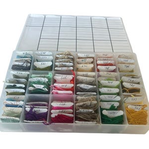 May include: A clear plastic storage container with a hinged lid featuring a grid pattern, filled with numerous small white cardboard bobbins wrapped in multi-colored embroidery floss. Each bobbin is labeled with a handwritten numerical code.