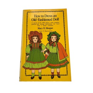 Op de afbeelding: Een geel paperbackboek getiteld How to Dress an Old-Fashioned Doll door Mary H. Morgan. De kaft heeft een zwarte decoratieve rand en een kleurrijke illustratie van twee poppen in groene jurken, mutsjes en een oranje cape.