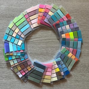 Puede incluir: Una disposición circular de blocs de notas adhesivas de colores. Los blocs son de varios tonos de rosa, azul, verde, amarillo y naranja. Cada bloc está en una funda de plástico transparente. El fondo es una superficie de madera marrón claro.